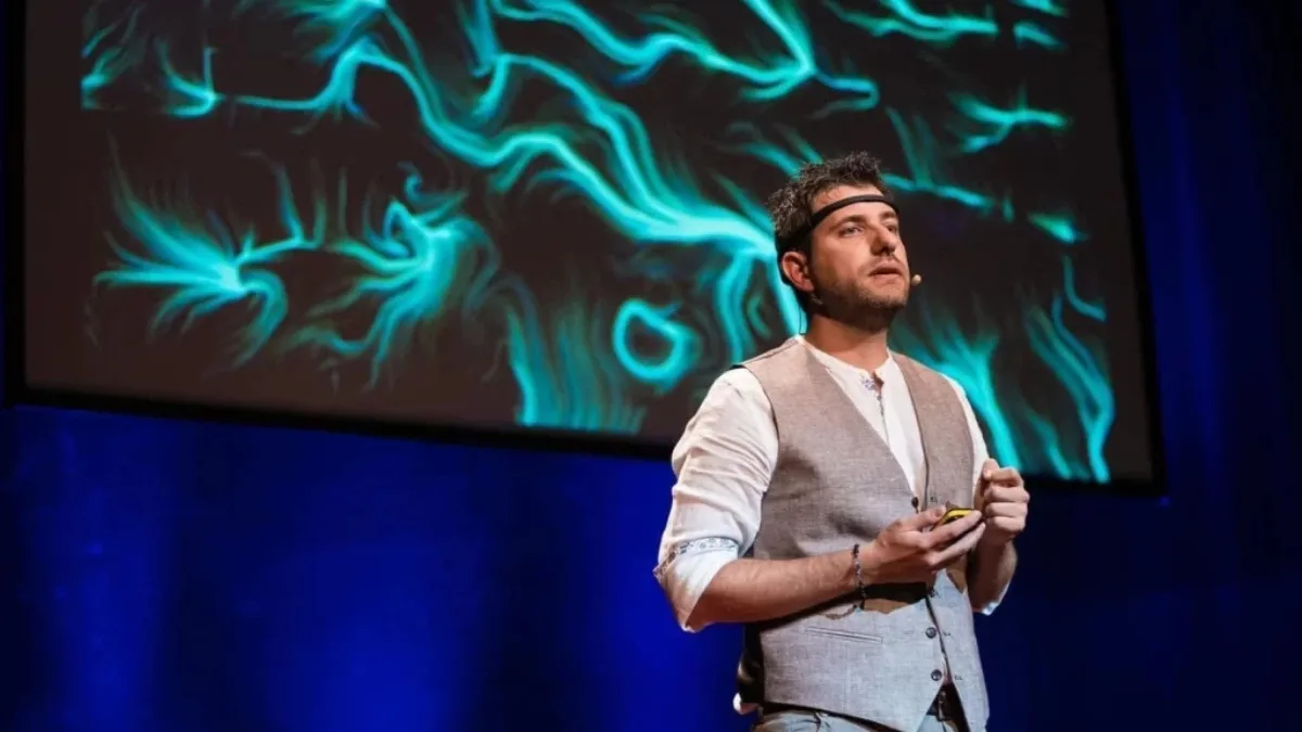 Un uomo parla sul palco di una conferenza, in piedi, con un sensore EEG sulla fronte e un microfono ad archetto. Indossa una camicia chiara con gilet grigio e tiene in mano un telecomando. Alle sue spalle, su un grande schermo, è proiettata un'opera dalle fitte linee e dai vortici turchesi su fondo scuro. L'ambiente è in penombra con luci blu.