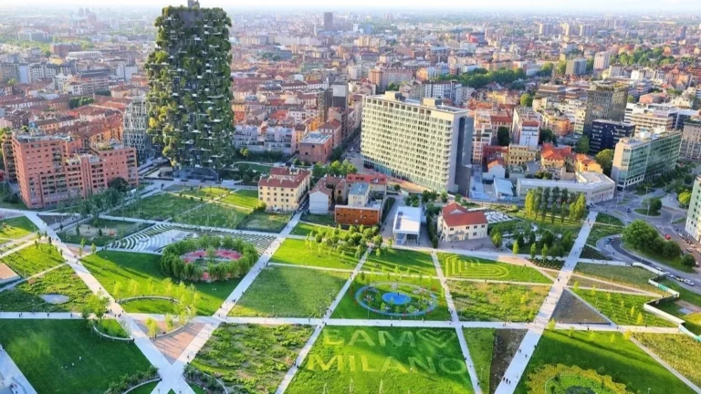 Veduta aerea del parco Biblioteca degli Alberi di Milano, con prati geometrici attraversati da vialetti diagonali, aiuole disegnate e alberi; sulla sinistra svetta un grattacielo interamente ricoperto di vegetazione, mentre sullo sfondo si estendono i tetti e gli edifici della città fino all'orizzonte.