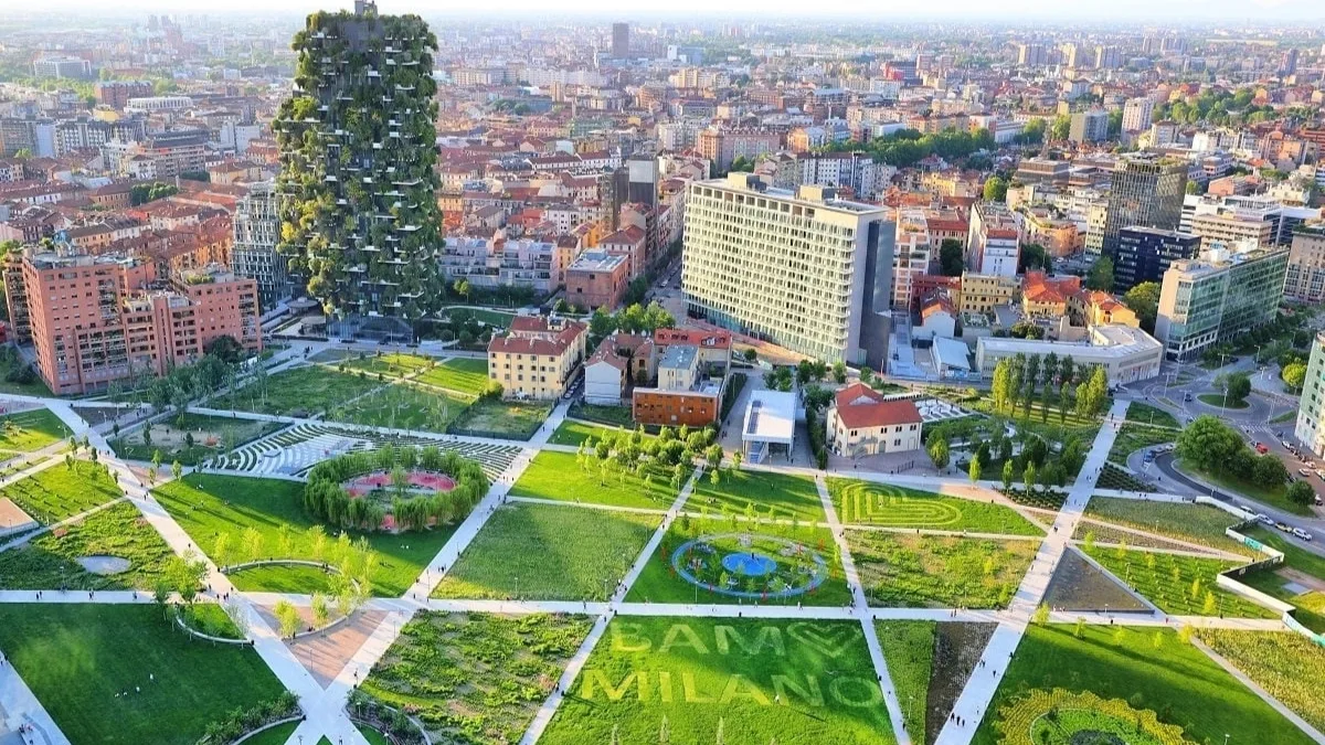 Veduta aerea del parco Biblioteca degli Alberi di Milano, con prati geometrici attraversati da vialetti diagonali, aiuole disegnate e alberi; sulla sinistra svetta un grattacielo interamente ricoperto di vegetazione, mentre sullo sfondo si estendono i tetti e gli edifici della città fino all'orizzonte.