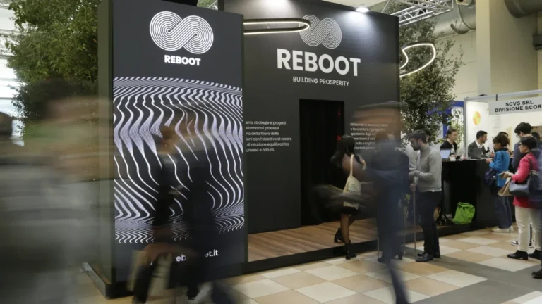 L'esterno di uno stand fieristico nero con il logo Reboot, un simbolo a nastro intrecciato, e la scritta "REBOOT BUILDING PROSPERITY"; sulla facciata di sinistra un pannello con fitte linee ondulate bianche su fondo scuro e alcuni testi descrittivi, mentre davanti alla struttura passano diversi visitatori mossi e, sulla destra, altre persone in piedi accanto a un banco.