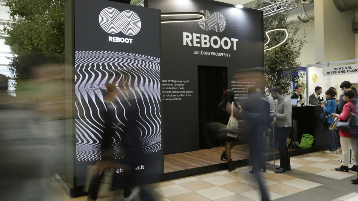 L'esterno di uno stand fieristico nero con il logo Reboot, un simbolo a nastro intrecciato, e la scritta "REBOOT BUILDING PROSPERITY"; sulla facciata di sinistra un pannello con fitte linee ondulate bianche su fondo scuro e alcuni testi descrittivi, mentre davanti alla struttura passano diversi visitatori mossi e, sulla destra, altre persone in piedi accanto a un banco.