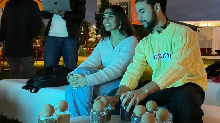Scena serale all'aperto: due persone sedute su una seduta bianca, una donna con maglione azzurro e mani intrecciate che sorride guardando di lato, e un uomo con maglione giallo illuminato che allunga una mano verso un tavolo. Sul tavolo diversi piccoli barattoli di vetro contenenti materiale granuloso e chiusi da sfere di sughero. L'uomo ha un sottile sensore appoggiato al collo. Dietro, una persona in piedi tiene un computer portatile aperto; sullo sfondo prato, strutture e luci dell'evento.