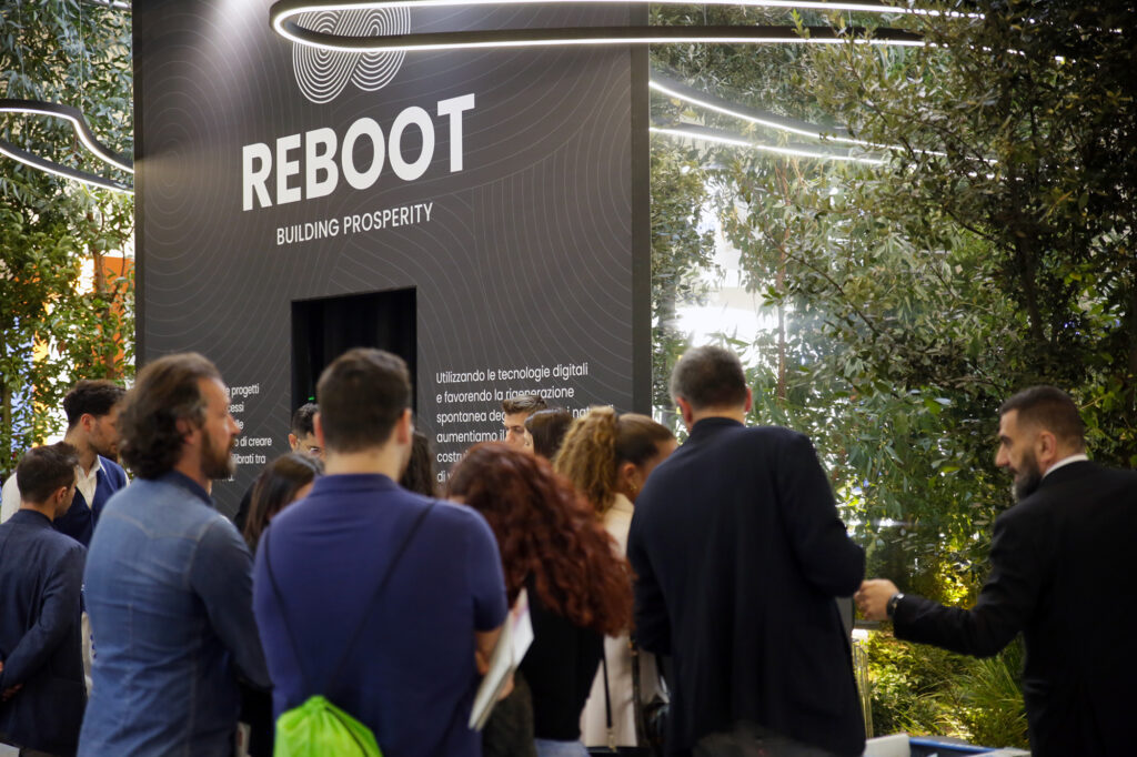 Una folla di persone radunata davanti allo stand Reboot con la scritta "Reboot – Building Prosperity" e un ingresso scuro; intorno piante e vegetazione.