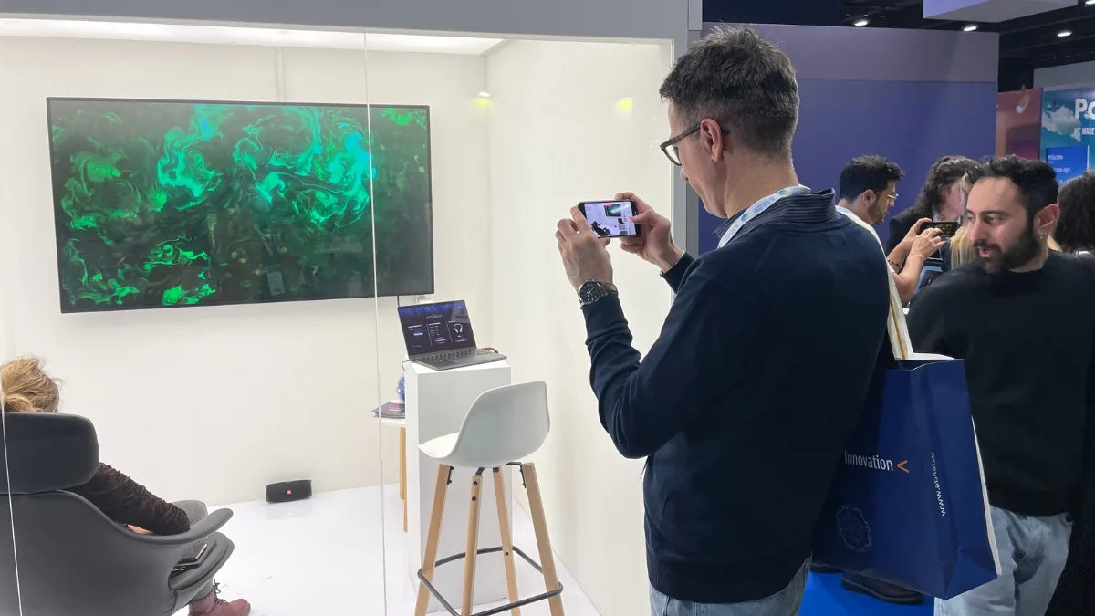 Uno stand fieristico dalle pareti bianche con un grande schermo a parete che mostra un'opera a volute verdi su fondo scuro; davanti allo schermo un uomo in maglione scuro fotografa il monitor con lo smartphone, mentre su un piano bianco è appoggiato un computer portatile con uno sgabello accanto e, sulla destra, altri visitatori con buste dell'evento.