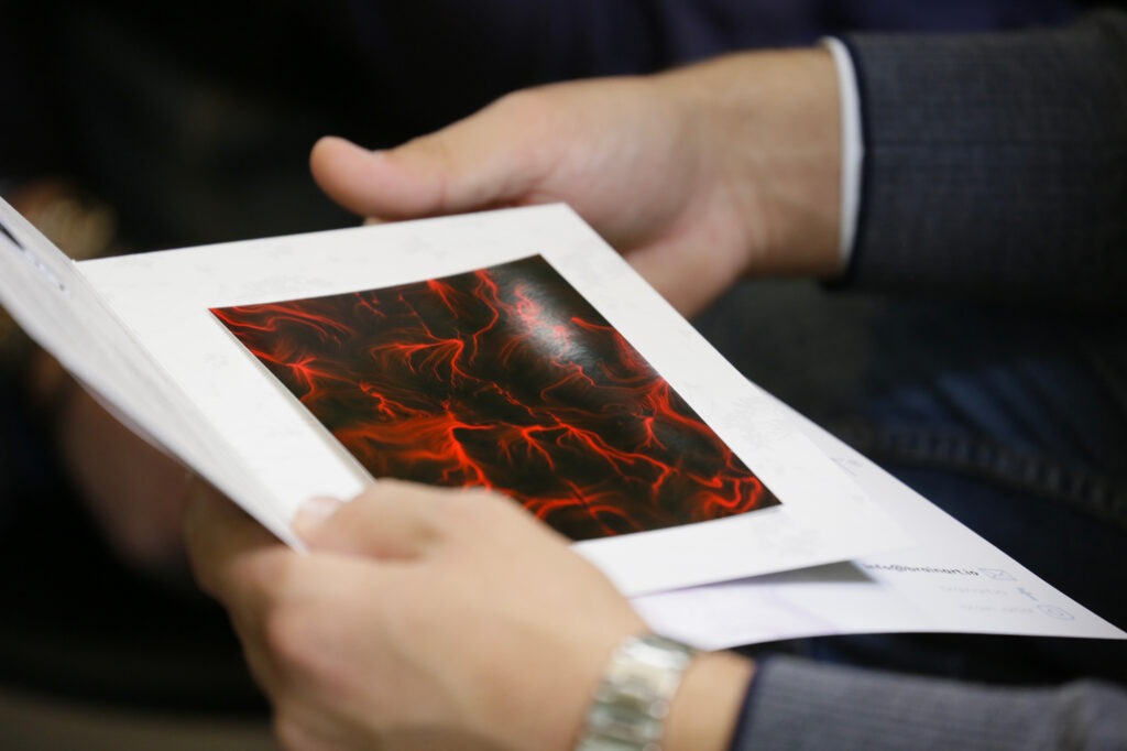 Le mani di un uomo tengono aperto un pieghevole BrainArt® con una stampa dell'opera dalle linee e dai vortici rossi su fondo scuro, incorniciata di bianco.