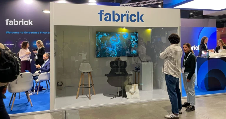 Lo stand Fabrick a una fiera, con il logo "fabrick" e una stanza dalle pareti vetrate che contiene uno schermo con un'opera BrainArt® dalle linee e dai vortici blu e una poltrona; accanto la parete "Welcome to Embedded Finance" con alcuni visitatori.