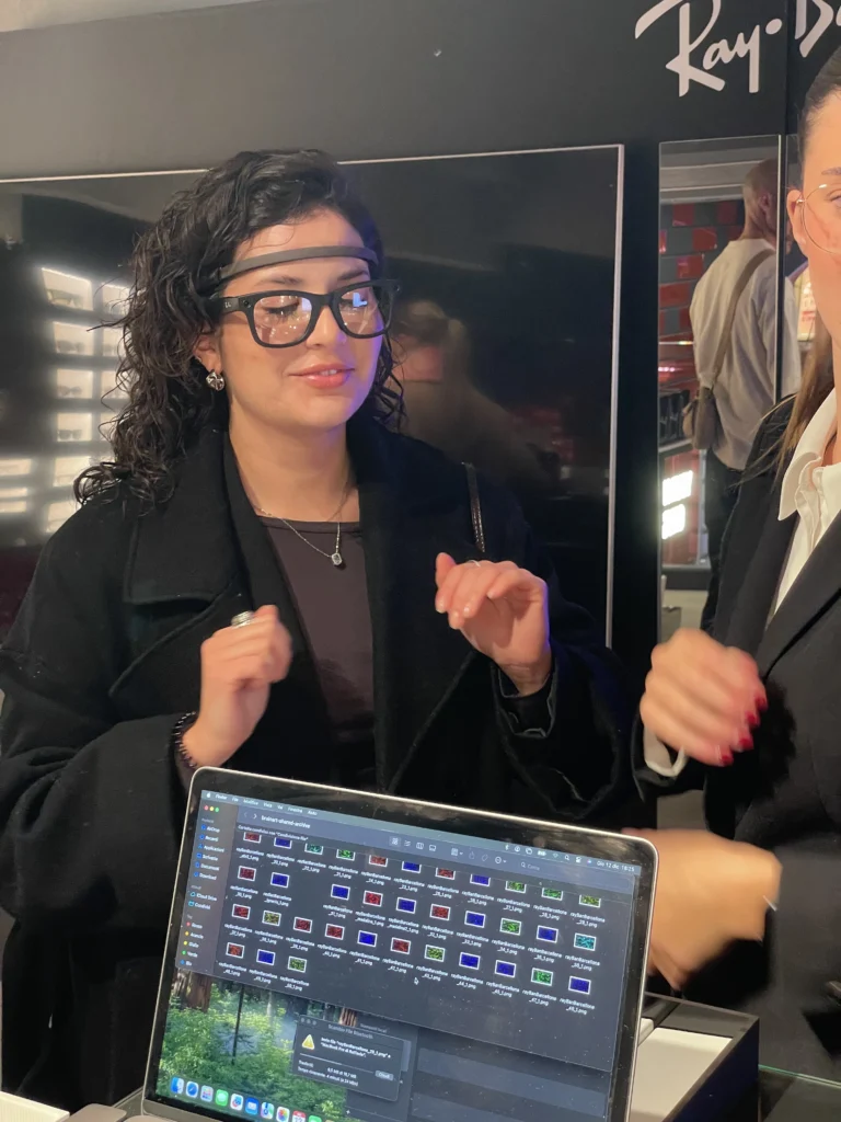 Una donna con un sensore EEG e occhiali Ray-Ban parla e gesticola davanti a un laptop che mostra una cartella con numerose miniature delle opere generate; accanto una donna dello staff, sullo sfondo il logo Ray-Ban.