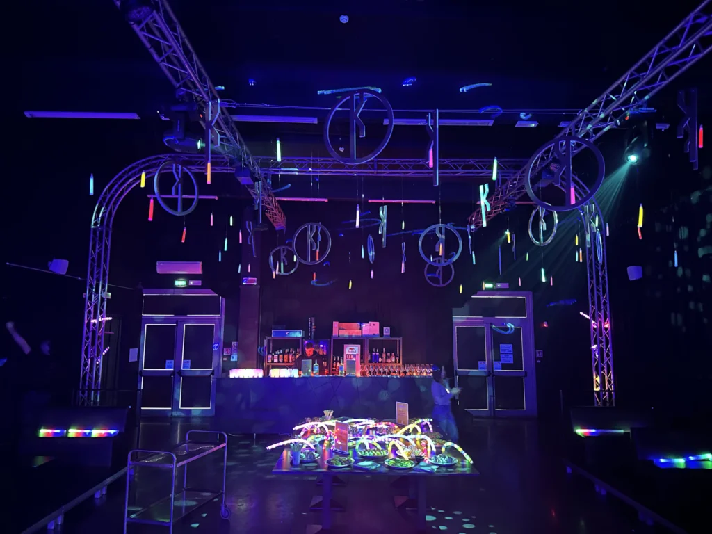 Lo spazio dell'evento al buio, con il bancone bar illuminato di viola, decorazioni circolari e tubi al neon appesi al soffitto e un tavolo con allestimenti luminosi colorati al centro.
