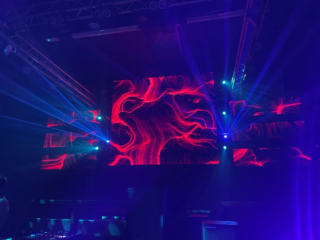 Un grande schermo mostra un'opera dalle linee e dai vortici rossi e arancioni su fondo scuro, circondato da fasci laser blu in uno spazio evento buio con strutture a soffitto.