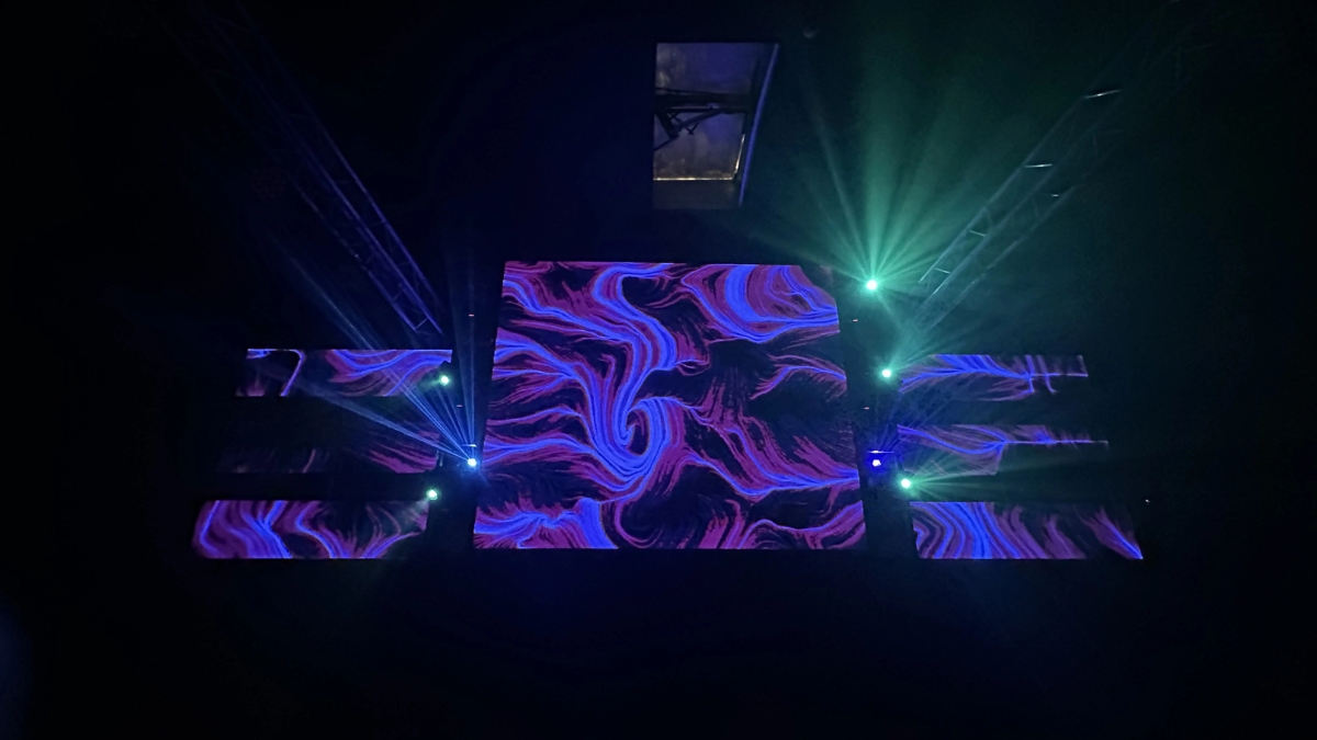 Uno spazio evento al buio con diversi schermi che mostrano opere dalle linee e dai vortici viola, rosa e blu, attraversato da fasci laser verdi e blu; in alto le strutture del soffitto.
