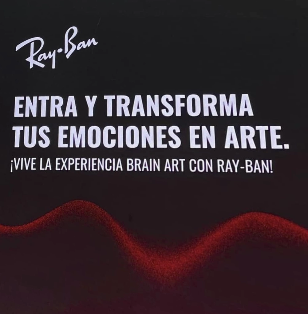 Cartello con il logo Ray-Ban su fondo scuro e la scritta in spagnolo "Entra y transforma tus emociones en arte. ¡Vive la experiencia Brain Art con Ray-Ban!", con un profilo ondulato rosso in basso.