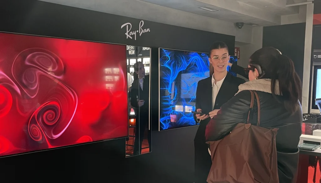 Due grandi schermi allo store Ray-Ban, sotto il logo del brand: uno mostra un effetto grafico rosso con volute, l'altro un'opera dalle linee e dai vortici blu; accanto una donna dello staff con un dispositivo in mano parla con una visitatrice che indossa un sensore EEG.