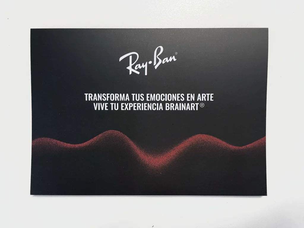 La copertina di un pieghevole nero con il logo Ray-Ban, la scritta in spagnolo "Transforma tus emociones en arte – Vive tu experiencia BrainArt®" e un profilo ondulato rosso.