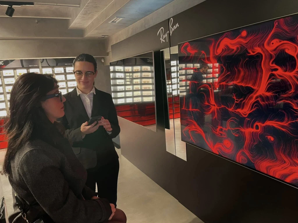 Una visitatrice con occhiali Ray-Ban e un sensore EEG osserva un grande schermo che mostra un'opera dalle linee e dai vortici rossi su fondo nero, accompagnata da una donna dello staff con un dispositivo in mano; sullo sfondo le vetrine con gli occhiali esposti.