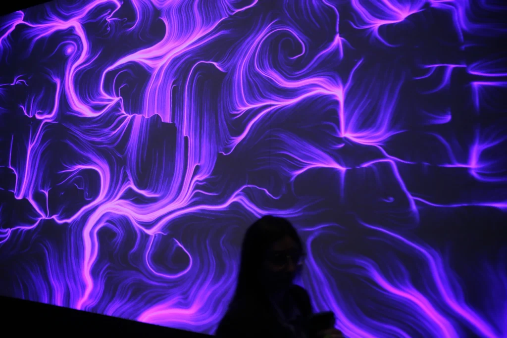 Una proiezione a grande formato occupa l'intera inquadratura con fitte linee e vortici viola e rosa su fondo scuro; in basso a destra si staglia in controluce la sagoma di una persona di profilo, con i capelli lunghi e il volto in ombra.