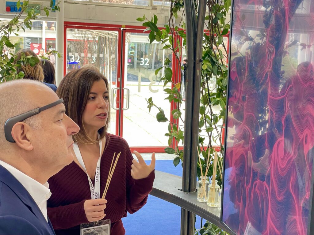 Un uomo di profilo con una fascia nera sulla fronte osserva un grande schermo verticale che mostra fitte linee rosa e magenta; accanto a lui una donna con badge Cosmoprof al collo tiene in mano dei bastoncini di legno e indica lo schermo, mentre sullo sfondo si vedono le vetrate del padiglione fieristico e due boccette con bastoncini appoggiate su una mensola.