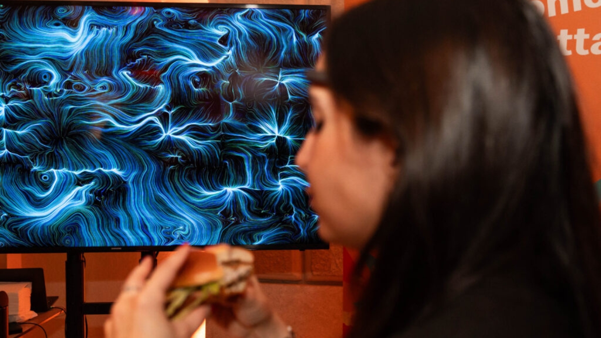 Una donna in primo piano, sfocata, porta alla bocca un panino davanti a un grande schermo che mostra un'opera dalle linee e dai vortici blu e azzurri su fondo scuro; sullo sfondo una parete arancione.