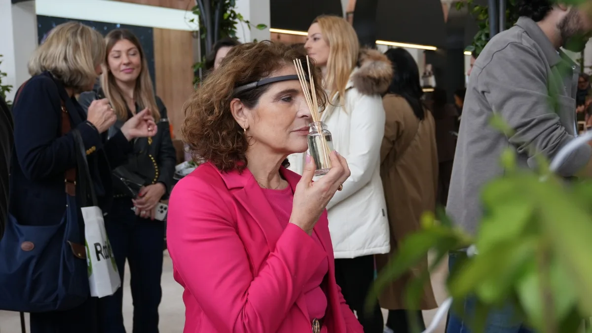 Una donna con capelli ricci e giacca rosa, con una fascia nera sulla fronte, avvicina al viso una boccetta di vetro con bastoncini di legno e tiene lo sguardo abbassato; alle sue spalle, altri visitatori in piedi all'interno di uno stand fieristico con piante verdi.