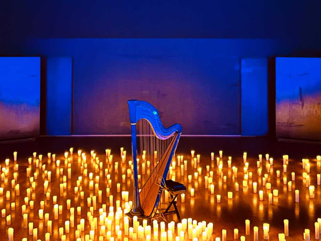 Un'arpa blu con lo sgabello vuoto al centro di un pavimento coperto da centinaia di candele accese, in una sala illuminata di blu con pannelli chiari alle pareti.