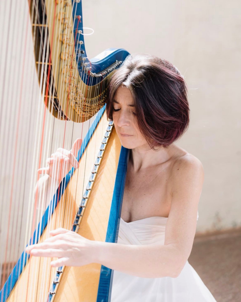 Primo piano di una musicista in abito bianco senza spalline che suona un'arpa dalla colonna blu e dalla tavola armonica in legno chiaro; ha gli occhi socchiusi, il capo inclinato verso lo strumento e le mani sulle corde, davanti a una parete chiara illuminata dal sole.