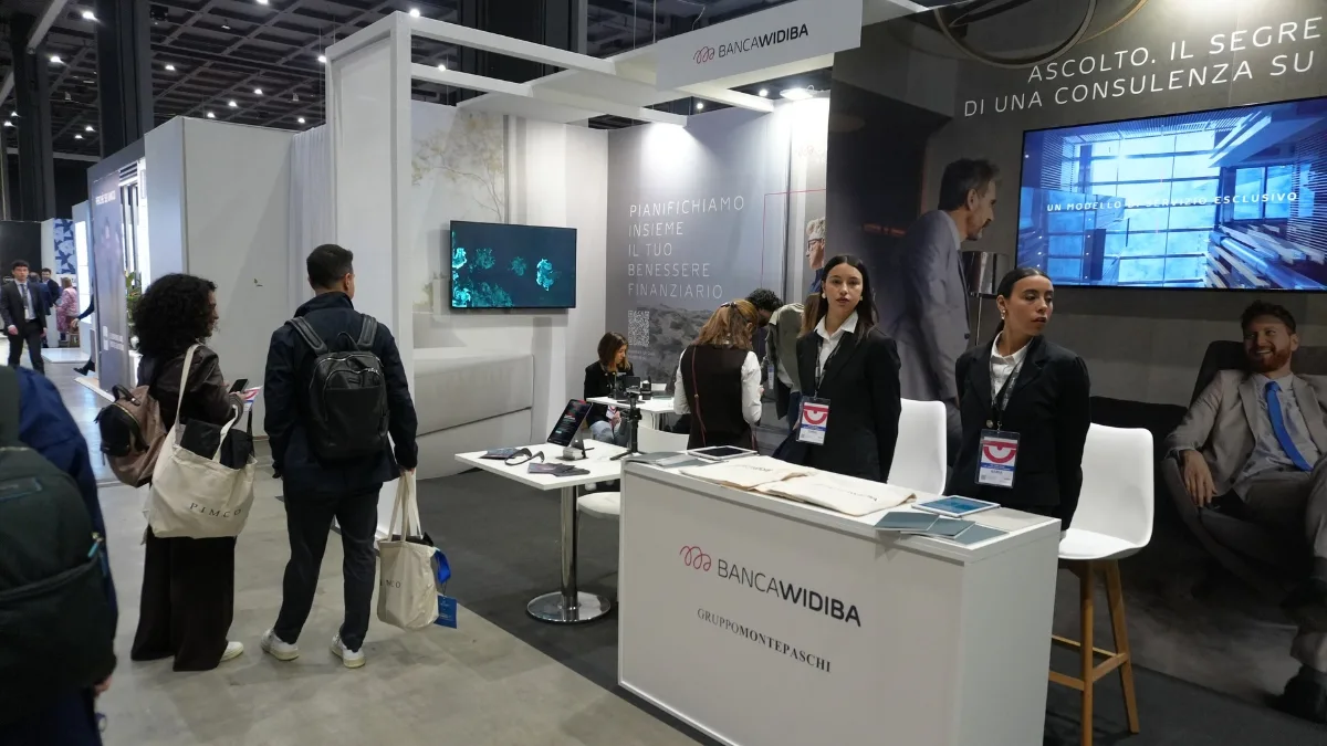 Lo stand fieristico di Banca Widiba, con il logo sulla trave superiore e sul bancone d'accoglienza insieme alla scritta Gruppo Montepaschi; sulla parete di sinistra uno schermo mostra forme turchesi su fondo nero accanto alla scritta "Pianifichiamo insieme il tuo benessere finanziario", mentre a destra un grande schermo con un uomo in giacca affianca due hostess in blazer scuro con badge; in primo piano alcuni visitatori di spalle con borse di tela e zaini.