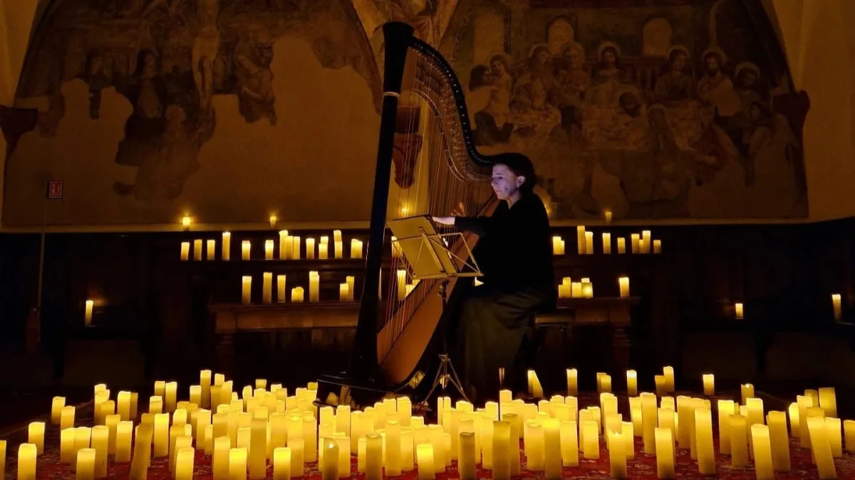 Una musicista vestita di scuro suona un'arpa davanti a un leggio illuminato, seduta al centro di una sala storica con due lunette affrescate alle pareti; il pavimento davanti a lei è coperto da decine di candele accese disposte su un tappeto.