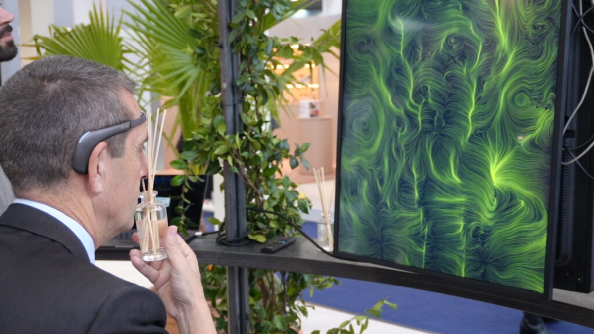 Un uomo con un sensore EEG avvicina al viso un diffusore di vetro con i bastoncini profumati, davanti a un monitor verticale che mostra un'opera dalle linee e dai vortici verdi; intorno le piante dell'allestimento.