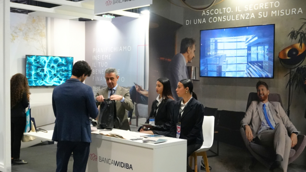 Lo stand di Banca Widiba a una fiera, con il logo del brand, il messaggio "Ascolto. Il segreto di una consulenza su misura" e uno schermo che mostra un'opera BrainArt® dalle linee e dai vortici blu; al banco alcune hostess e alcuni visitatori.