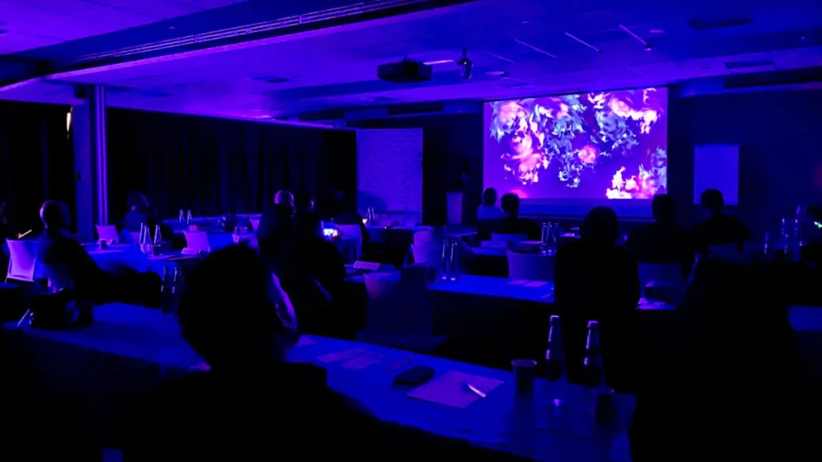 Una sala conferenze in penombra, illuminata di blu e viola, con file di tavoli e persone sedute di spalle. Sul grande schermo a parete è proiettata un'opera astratta dalle volute e dai vortici in tonalità viola, rosa e chiare su fondo scuro. Al centro del soffitto un videoproiettore; sui tavoli bottiglie, bicchieri e fogli.