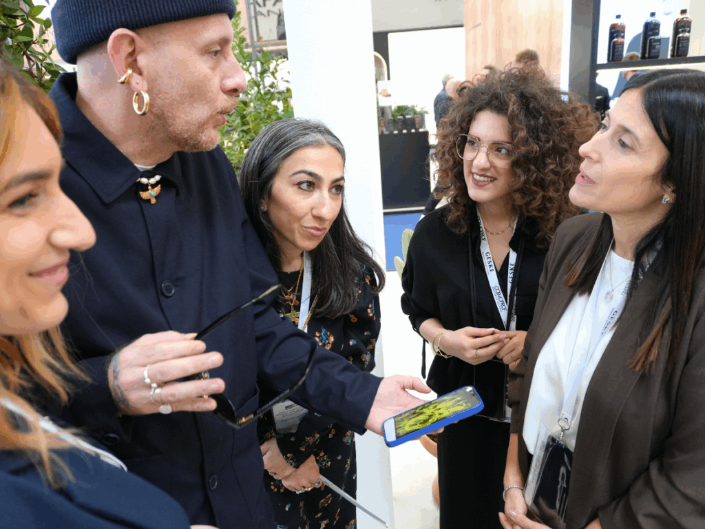 Un gruppo di persone conversa da vicino a uno stand fieristico. Un uomo con berretto scuro e orecchini tiene in mano uno smartphone con cover blu, sul cui schermo si vede un'opera dalle linee e dai vortici verdi, e lo mostra alle persone accanto. Intorno a lui alcune donne in dialogo, con badge al collo su cui si leggono le scritte "Cosmoprof" e "Geske". Sullo sfondo scaffali con flaconi di prodotti.