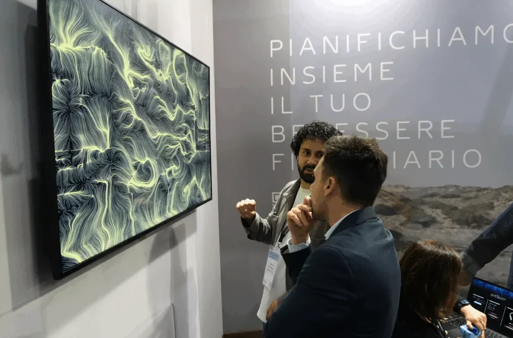 In uno stand fieristico, un uomo in giacca scura di profilo, con la mano al mento, e un altro uomo con giacca chiara che indica con il pugno osservano uno schermo a parete. Sullo schermo un'opera dalle fitte linee e dai vortici in tonalità verdi e dorate chiare su fondo scuro. Sulla parete di destra la scritta "Pianifichiamo insieme il tuo benessere finanziario"; a destra una persona seduta a un portatile.