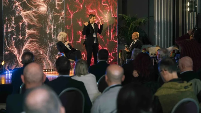 Immagine divisa in due scene di uno stesso evento. A sinistra, sul palco, uno speaker in piedi con un sensore EEG sulla fronte parla accanto a una relatrice seduta; alle loro spalle un grande schermo LED mostra un'opera dalle fitte linee e dai vortici rossi su fondo scuro, con una fila di luci colorate ai piedi del palco. A destra, un uomo seduto parla al microfono. In primo piano, di spalle, la platea gremita.