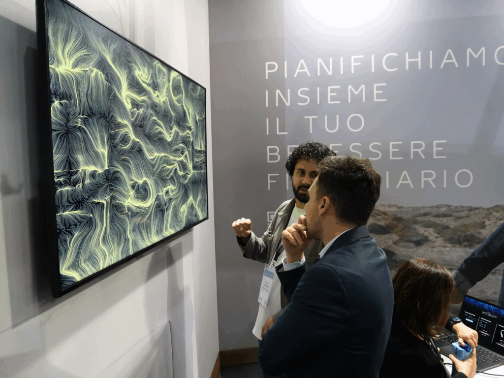 In uno stand fieristico, due uomini in dialogo ravvicinato osservano e commentano; uno indica con la mano verso uno schermo a parete sulla sinistra. Sullo schermo un'opera dalle fitte linee e dai vortici in tonalità verdi e dorate chiare su fondo scuro. Sulla parete di destra la scritta "Pianifichiamo insieme il tuo benessere finanziario". Uno dei due porta un pettorale al collo.