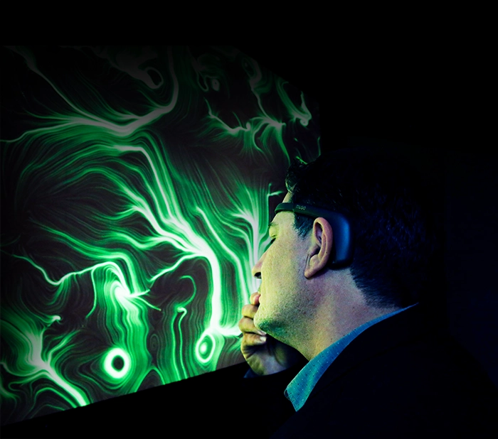 Uomo di profilo con un dispositivo EEG indossato sulla fronte osserva assorto un grande quadro BrainArt® dai vortici verdi su schermo, in un ambiente buio.