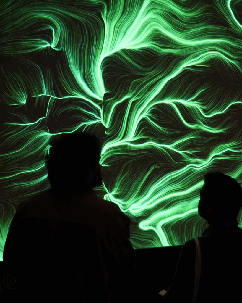 Due persone in silhouette osservano un grande quadro BrainArt® dalle linee verdi luminose proiettato su schermo, in un ambiente buio.