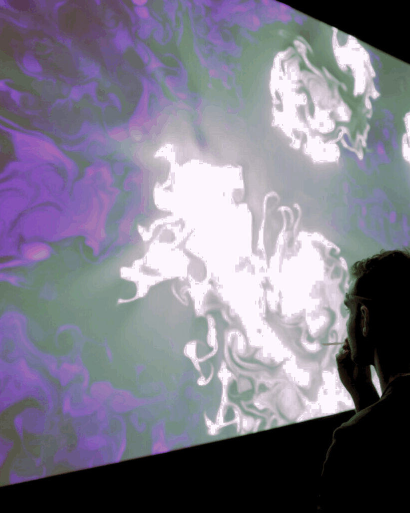 Persona in silhouette osserva un grande quadro BrainArt® dai vortici viola, verdi e bianchi proiettato su schermo, in un ambiente buio.