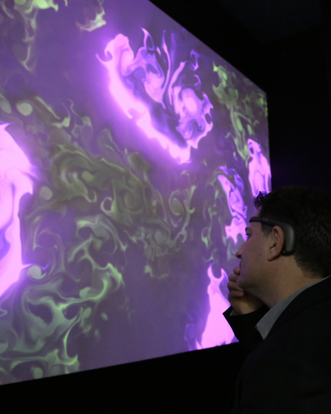 Uomo di profilo con un dispositivo EEG indossato sulla fronte osserva pensieroso un grande quadro BrainArt® dai vortici viola e verdi su schermo, in un ambiente buio.