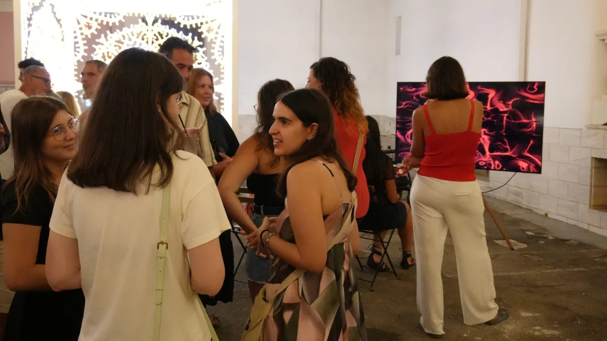 Un gruppo di persone in piedi conversa all'interno di una sala; sulla destra uno schermo appoggiato a terra mostra un'opera dalle linee e dai vortici rosa e magenta su fondo scuro, e una donna in canotta rossa vi si avvicina; sullo sfondo, pannelli decorativi bianchi traforati.