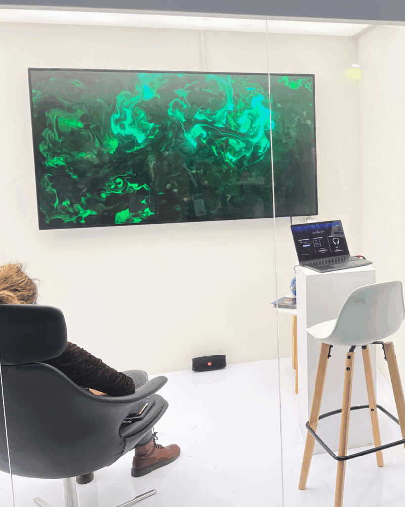 Persona seduta in poltrona in uno stand osserva un quadro BrainArt® verde su uno schermo a parete, accanto a un laptop posizionato su una postazione bianca.