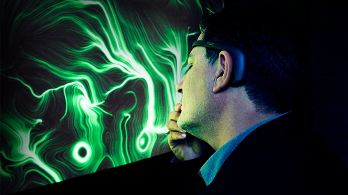 Un uomo di profilo, con un sensore EEG sulla testa e la mano vicino al mento, davanti a uno schermo che mostra un'opera dalle linee e dai vortici verdi su fondo nero.