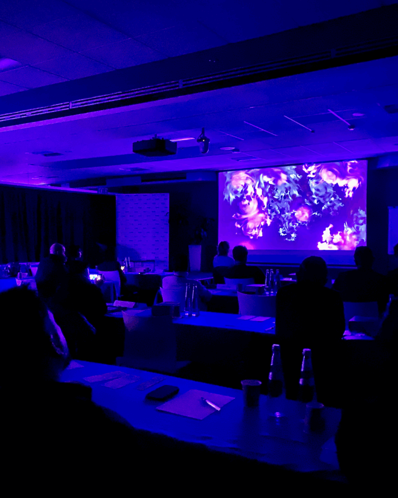 Sala conferenze in penombra con luci blu, un pubblico seduto ai tavoli osserva un quadro BrainArt® proiettato su grande schermo.