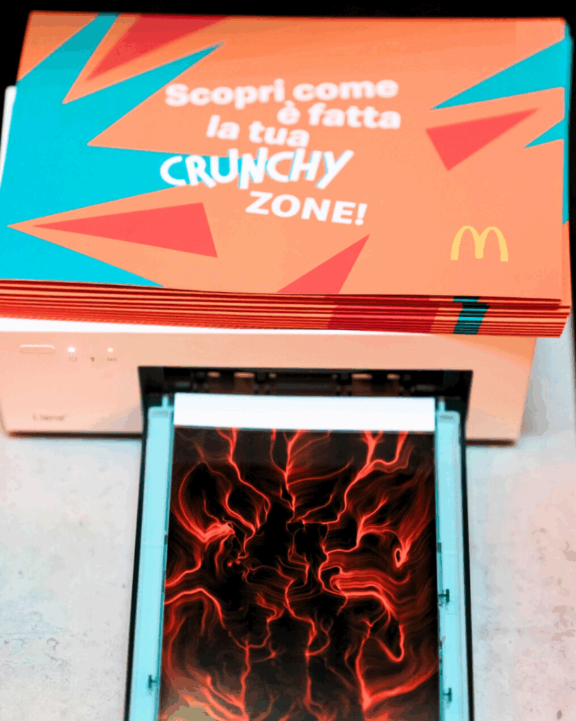 Una stampante produce un quadro BrainArt® dalle linee rosse, sopra una pila di pieghevoli McDonald's con la scritta "Scopri come è fatta la tua Crunchy Zone!"