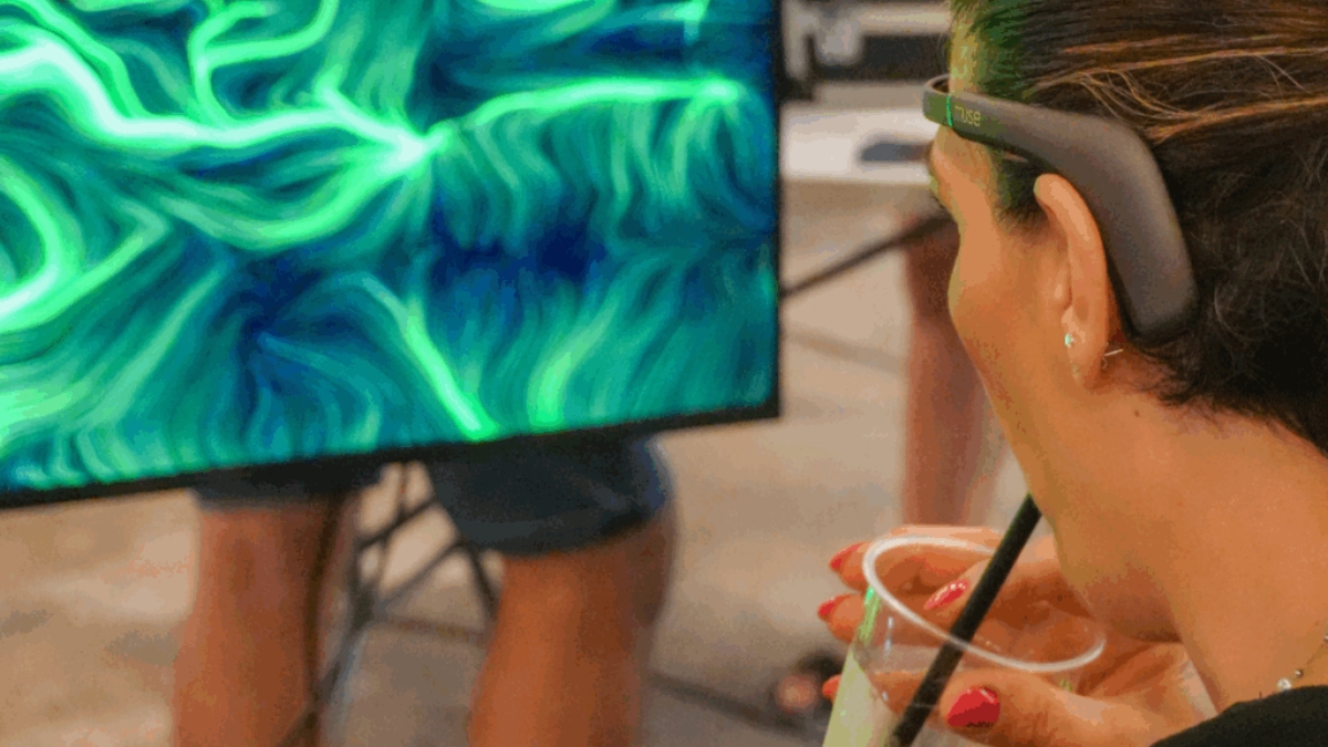 Donna di spalle con un dispositivo EEG indossato osserva un quadro BrainArt® verde su uno schermo, tenendo in mano un drink