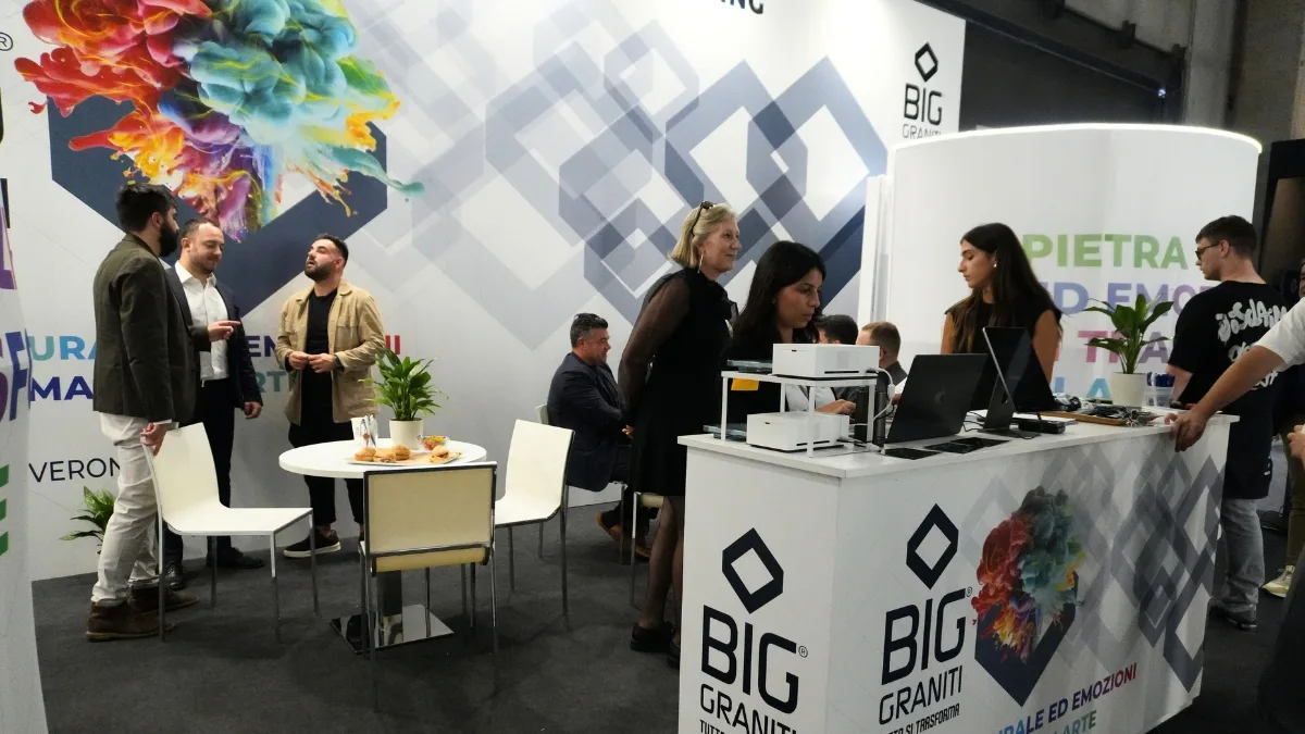 Stand fieristico con il logo BIG Graniti ripetuto sul bancone e sui pannelli di fondo, accanto a un'illustrazione di un cervello composto da schizzi di colore; alcune persone conversano in piedi attorno a un tavolino con dei vassoi, altre sono al bancone con computer portatili e stampanti.