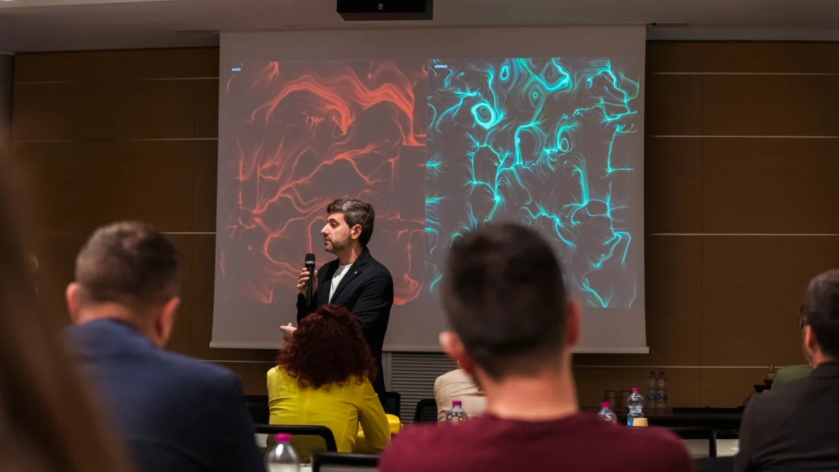In una sala meeting, un uomo in giacca scura parla al microfono davanti a un grande schermo. Sullo schermo due opere affiancate: a sinistra volute e vortici rossi, a destra fitte linee e vortici turchesi, entrambe su fondo scuro, ciascuna sormontata da un nome ("sonia" e "stefano"). In primo piano, di spalle, il pubblico seduto ai tavoli con bottiglie d'acqua.