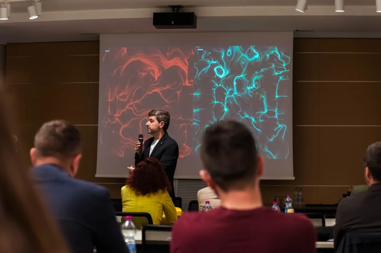 In una sala meeting, un uomo in giacca scura parla al microfono davanti a un grande schermo. Sullo schermo due opere affiancate: a sinistra volute e vortici rossi, a destra fitte linee e vortici turchesi, entrambe su fondo scuro, ciascuna sormontata da un nome ("sonia" e "stefano"). In primo piano, di spalle, il pubblico seduto ai tavoli con bottiglie d'acqua.