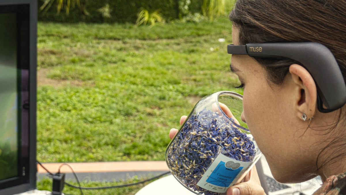 Primo piano di una donna ripresa di profilo da dietro, con un sensore EEG scuro indossato sulla fronte e sopra le orecchie. Avvicina al naso un barattolo di vetro pieno di fiori e petali essiccati azzurri e violacei, con un'etichetta sul fronte. A sinistra, sfocati, uno schermo scuro su una struttura bianca, cavi a terra, un prato verde e una parete vegetale.