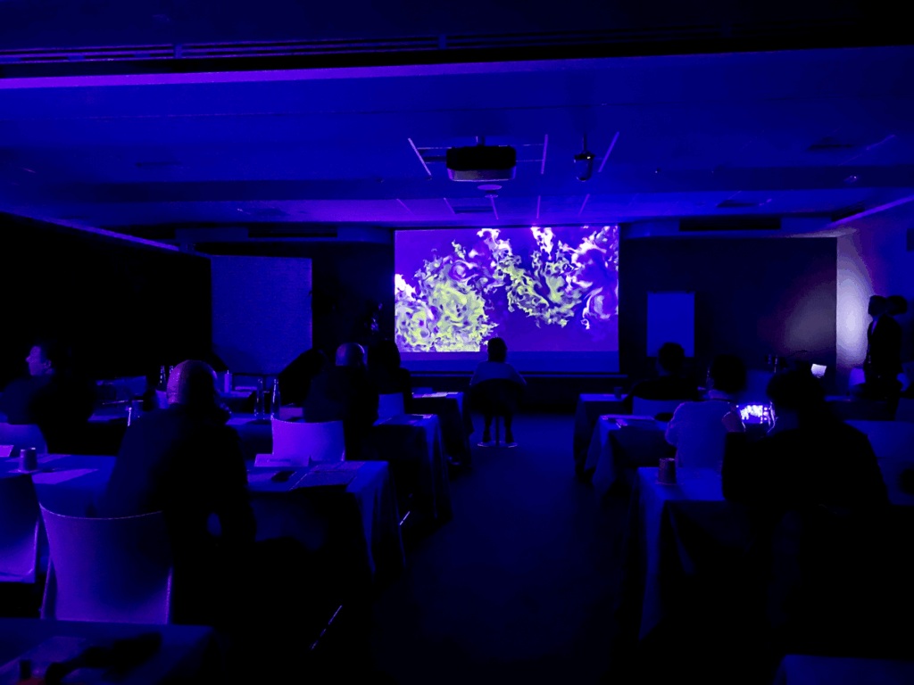 Una sala meeting in penombra, immersa in una luce blu e viola, con tavoli disposti su più file e persone sedute di spalle. Sullo schermo a parete è proiettata un'opera dalle volute e dai vortici verdi e lime su fondo scuro. Al centro del soffitto un videoproiettore; sui tavoli fogli, bicchieri e piccole luci.