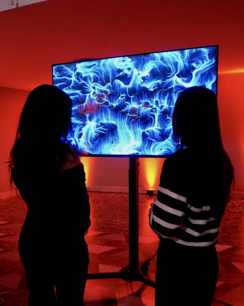 Un monitor su piantana mostra un'opera dalle fitte linee e dai vortici azzurri e bianchi su fondo scuro; davanti allo schermo, in controluce, si stagliano le sagome di due persone che lo osservano, in una sala illuminata da luci rosse e arancioni.
