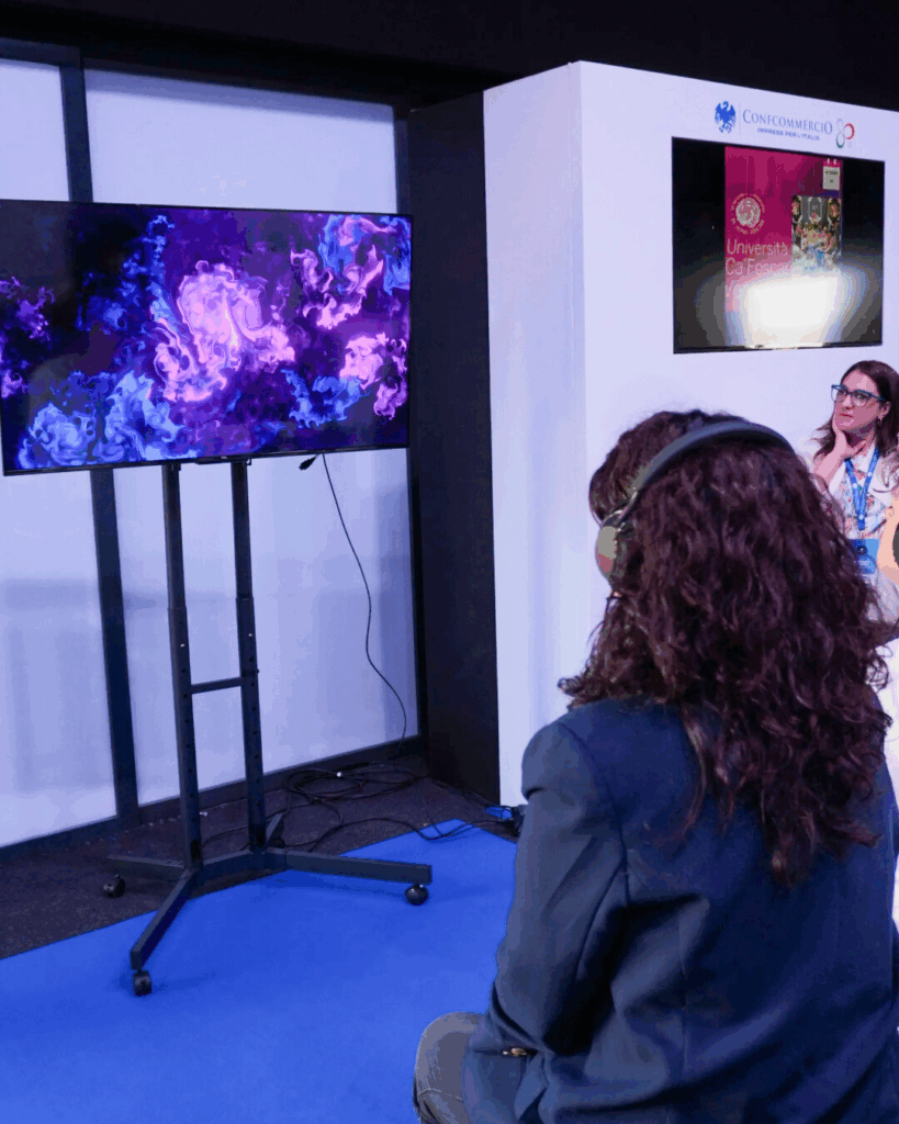 Una donna ripresa di spalle, con le cuffie, osserva un grande schermo su piedistallo che mostra un'opera dalle linee e dai vortici viola con un centro luminoso; accanto lo stand con il logo Confcommercio e un'altra persona.